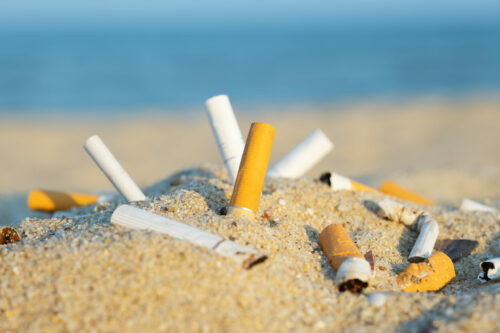 Cigarette butts in yellow sand on sea beach on coast against background of blue sky and sea. Problem of humanity. Cigarette smoking, bad habit. Nicotine addiction. Garbage Cigarette butts in yellow sand on sea beach on coast against background of blue sky and sea. Problem of humanity. Cigarette smoking, bad habit. Nicotine addiction. Garbage